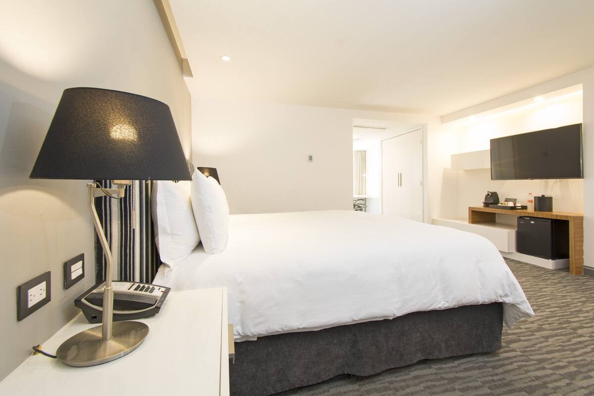 Ein Hotelzimmer mit einem großen Bett, einem Schreibtisch mit Lampe, einem Flachbildfernseher und einer kleinen Küchenzeile.