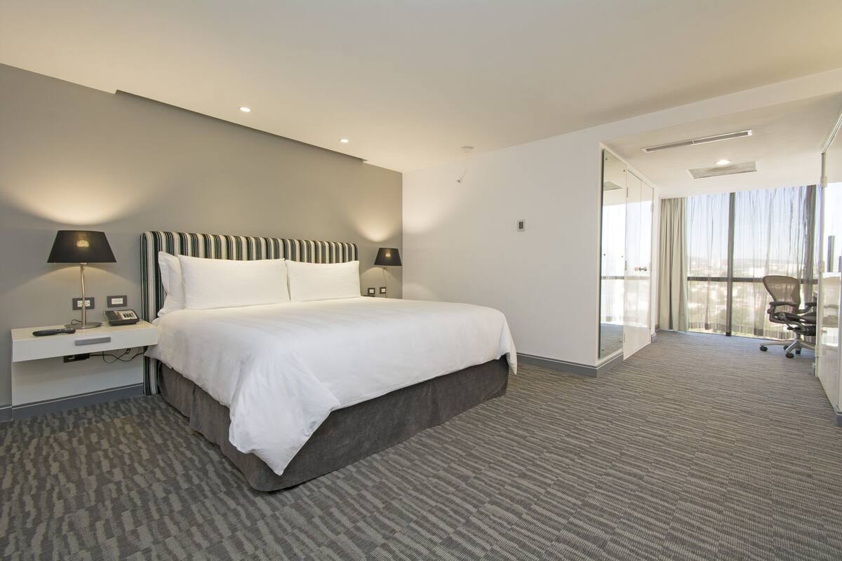 Ein Hotelzimmer mit einem großen Bett, Nachttischlampen, einem Schreibtisch mit Telefon und Blick auf die Stadt durch große Fenster.