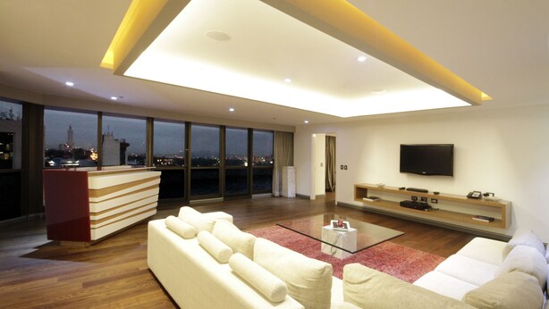 A modern living room with a high ceiling, a flat-screen TV mounted on the wall, a large window with a city view, and a red accent rug. - Presidente InterContinental Guadalajara by IHG (Zapopan)