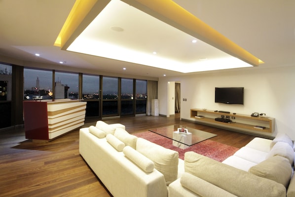 Un salón moderno con techo alto, un televisor de pantalla plana en la pared, un ventanal con vista a la ciudad y un tapete rojo decorativo.