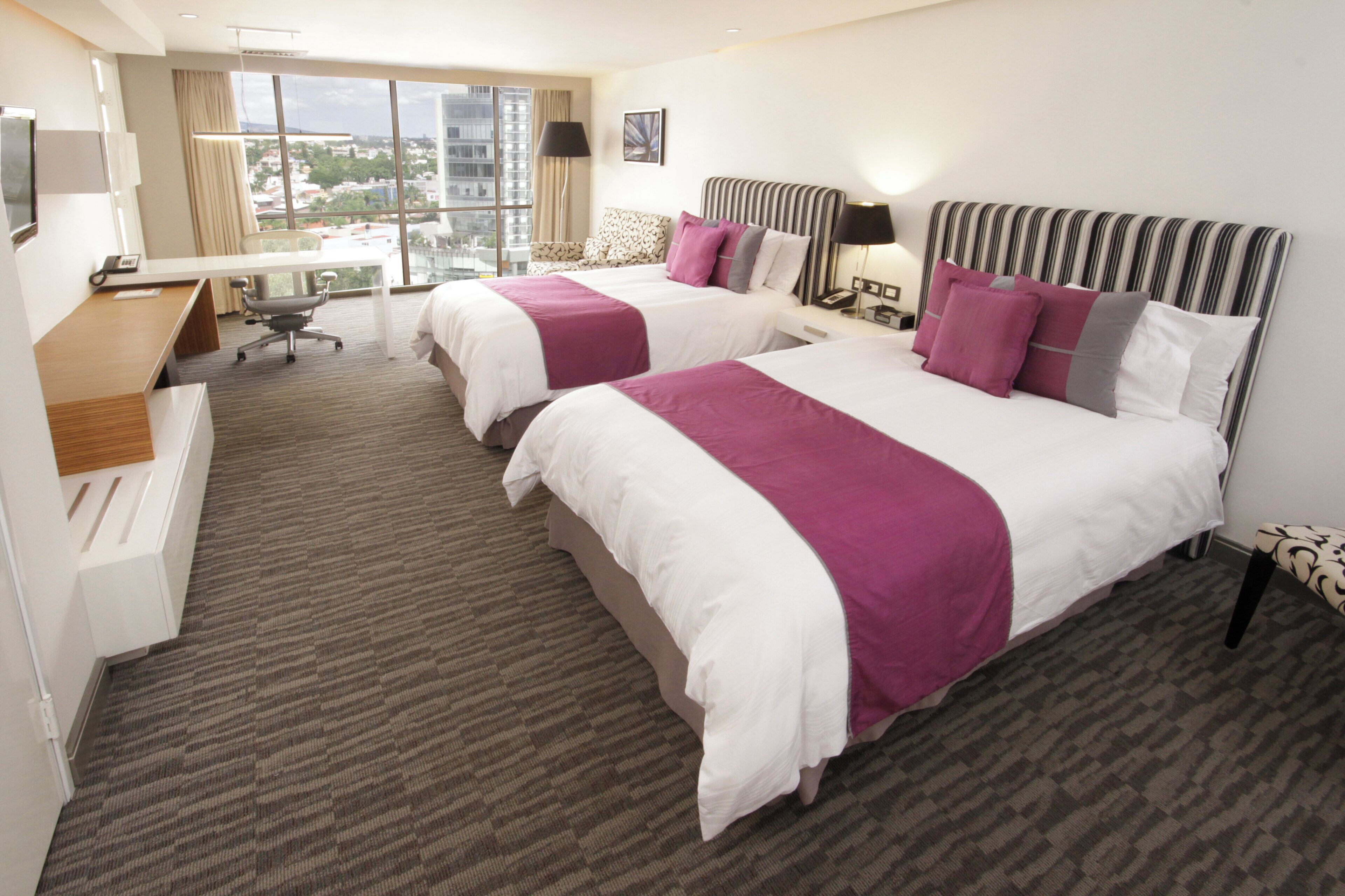 A hotel room with two beds, a desk, and a view of the city.