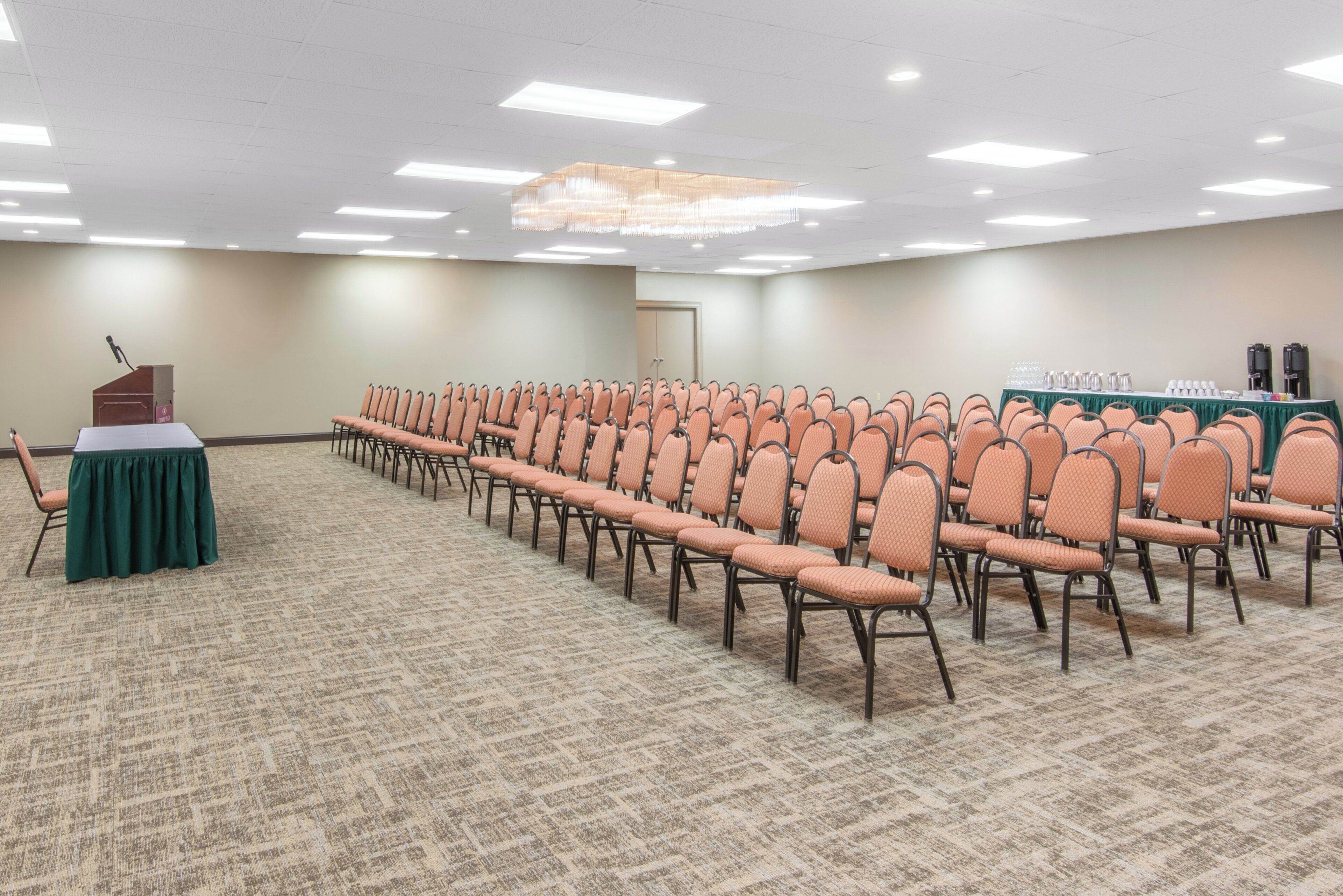 Sala de conferências com fileiras de cadeiras, um pódio e uma mesa com dispensadores de café e água.