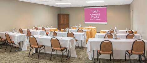 Un salón de conferencias con varias mesas, sillas y una pantalla de proyección mostrando 'CROWN PLAZA HOTELS & RESORTS'.