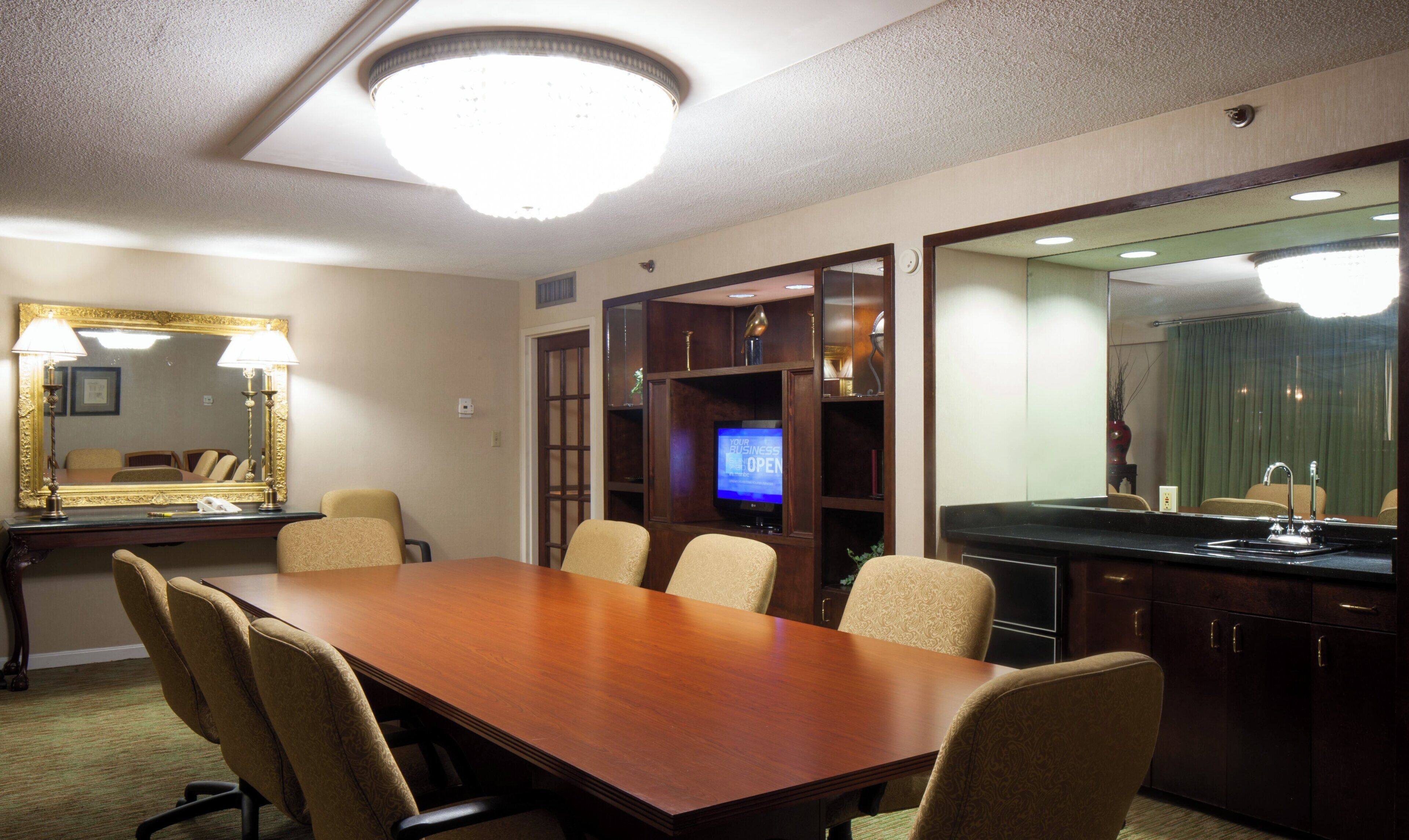 Una sala de conferencias con una mesa de madera, sillas color beige, un espejo grande, un televisor y una zona de kitchenette.