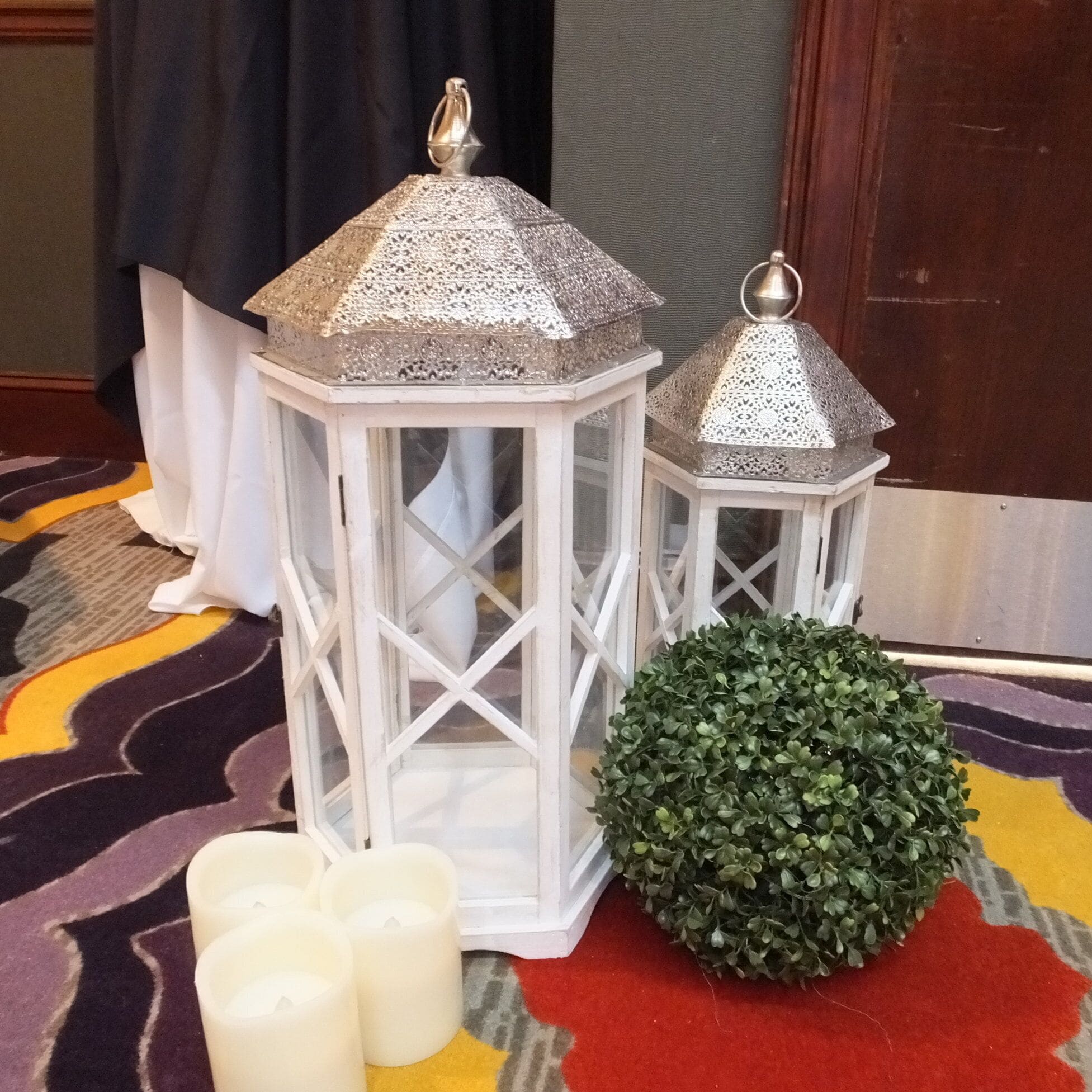 Two white lanterns with intricate metal tops, a green spherical plant, and several white candles on a patterned carpet.