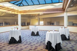 Un salón de conferencias con varias mesas, cada una con mantel blanco y fundas negras para sillas, y una alfombra estampada.