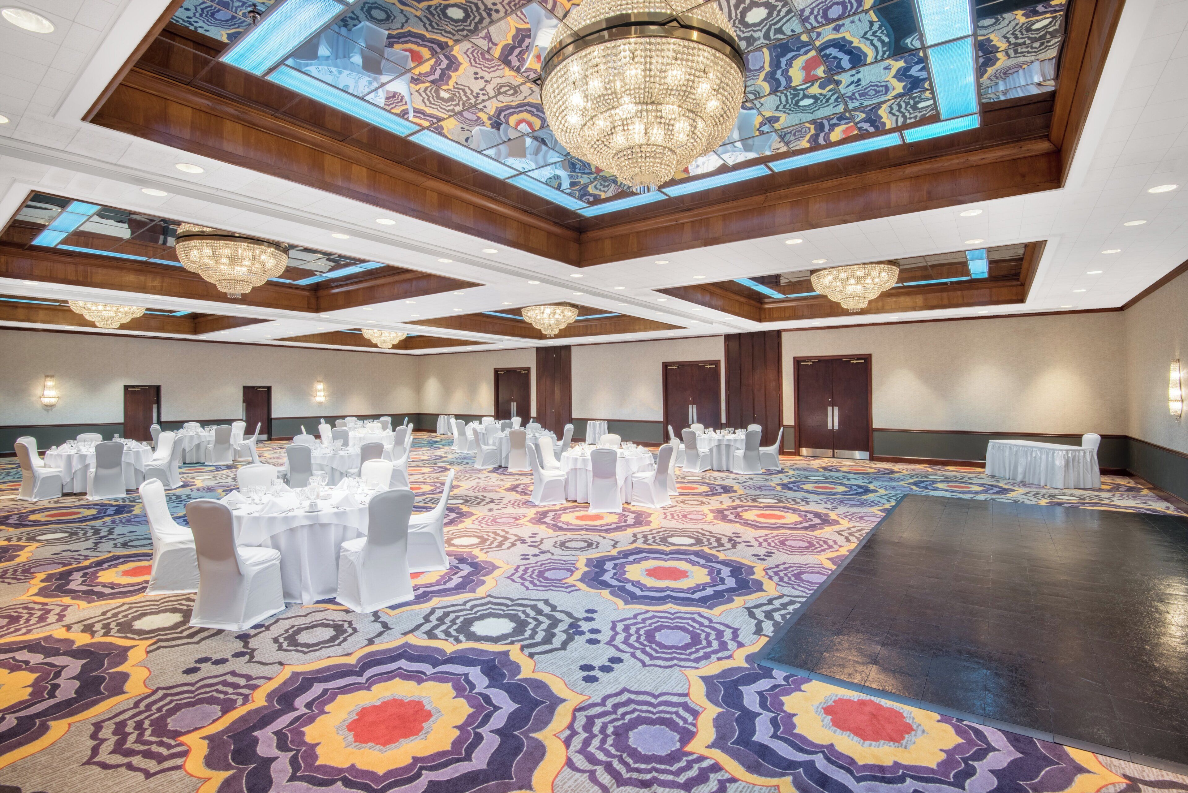 Une salle de banquet spacieuse, dotée d’un tapis à motifs, de nombreuses tables préparées pour des événements et de lustres décoratifs.