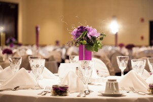 Una mesa de banquete dispuesta con cubertería de vidrio, un florero morado con flores y manteles blancos.