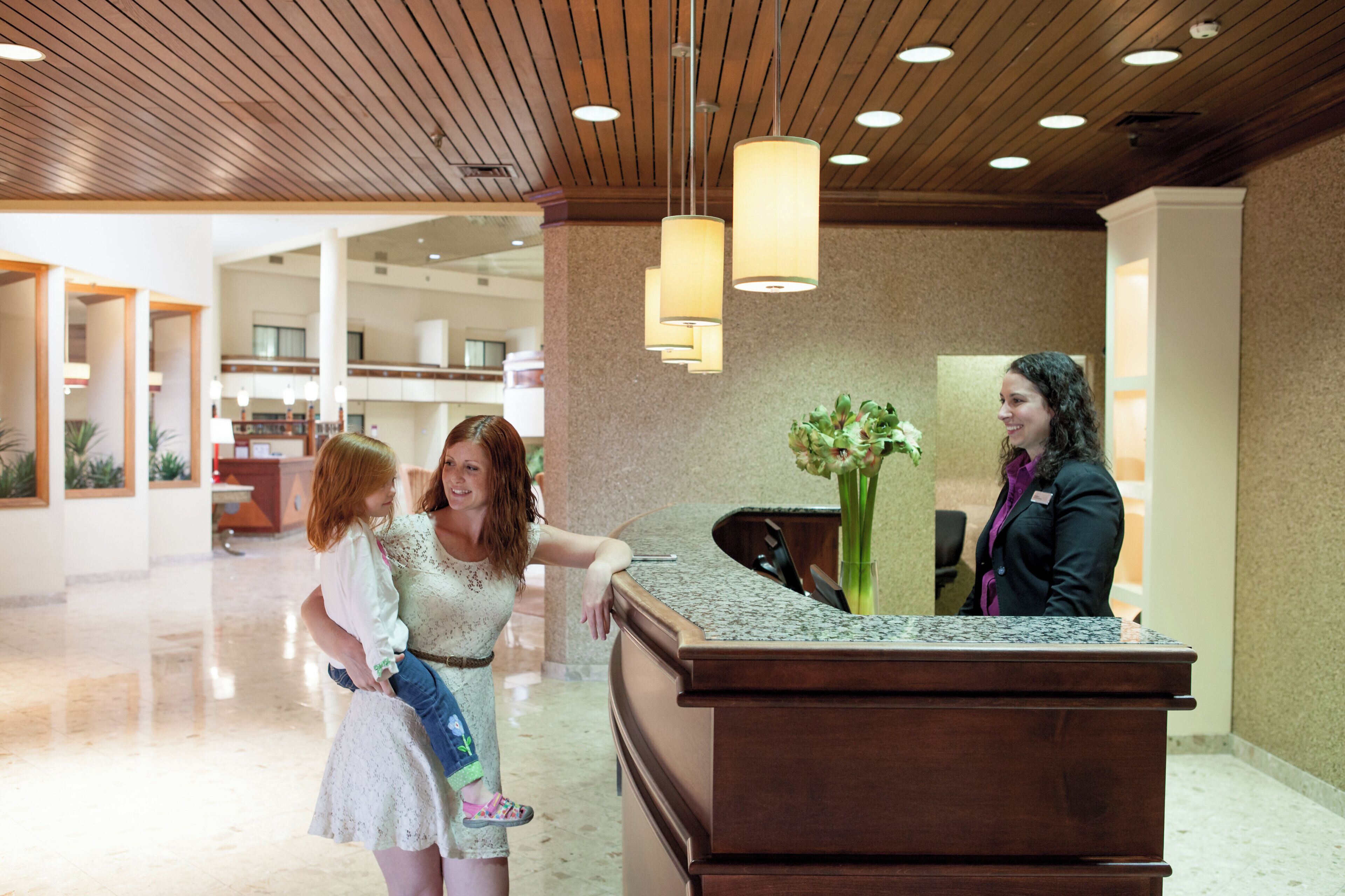 A reception desk in a hotel lobby with a modern design, including a wooden ceiling and pendant lighting.