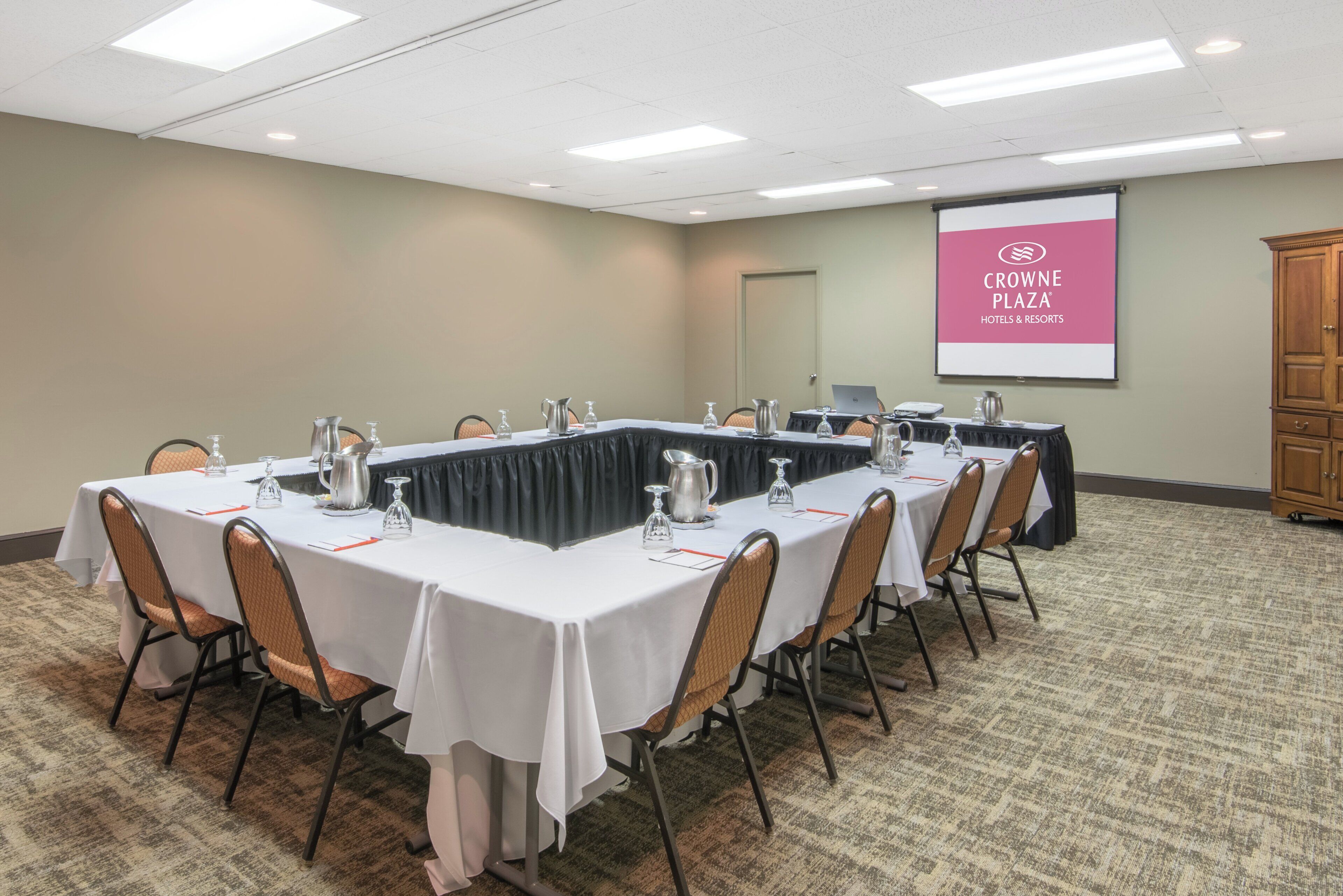 A conference room with multiple tables, chairs, and a projector screen displaying 'CROWNE PLAZA HOTELS & RESORTS'.