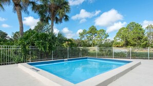 A rectangular swimming pool with a white border and a blue interior, surrounded by a white fence and lush greenery.