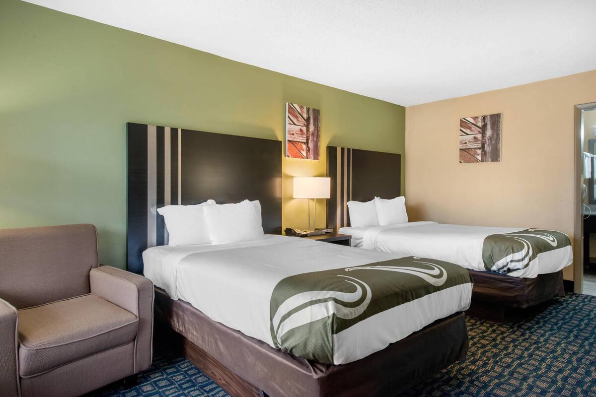 A hotel room with two beds, a chair, and a nightstand with a lamp.