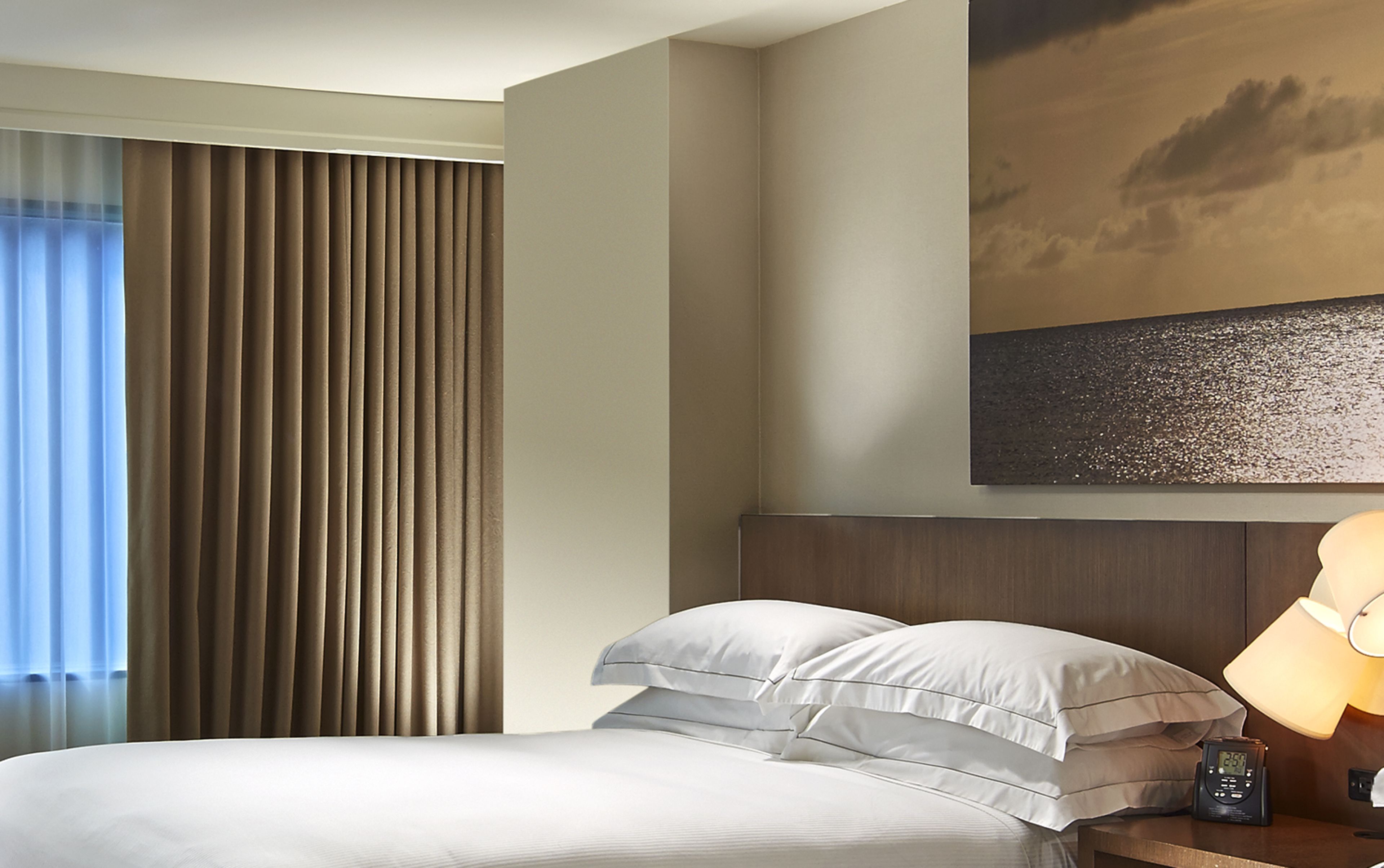 Una habitación de hotel con cama, cortinas, un cuadro y una mesita de noche con lámpara y reloj.