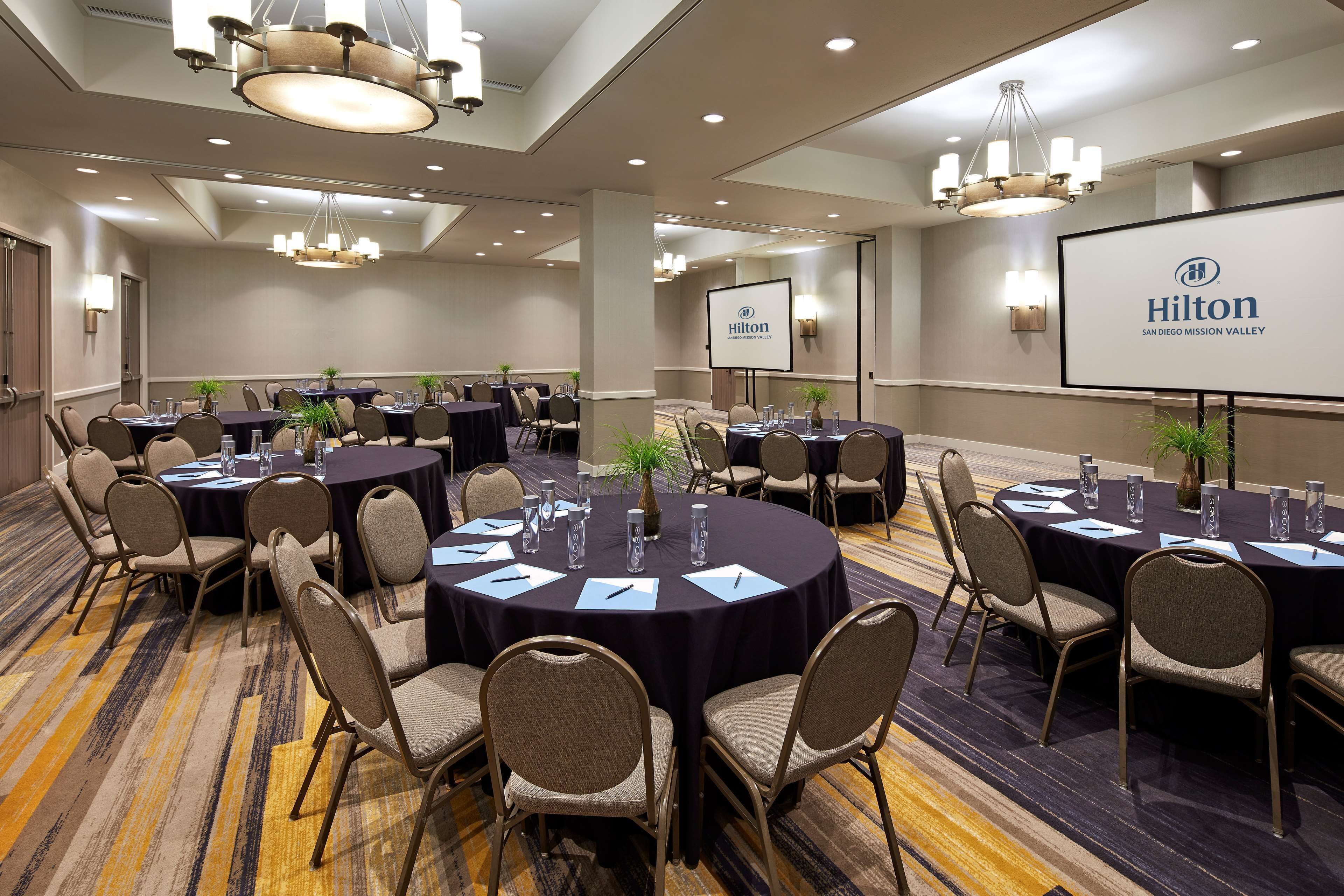 A conference room with multiple round tables, chairs, and a large screen displaying the Hilton logo.