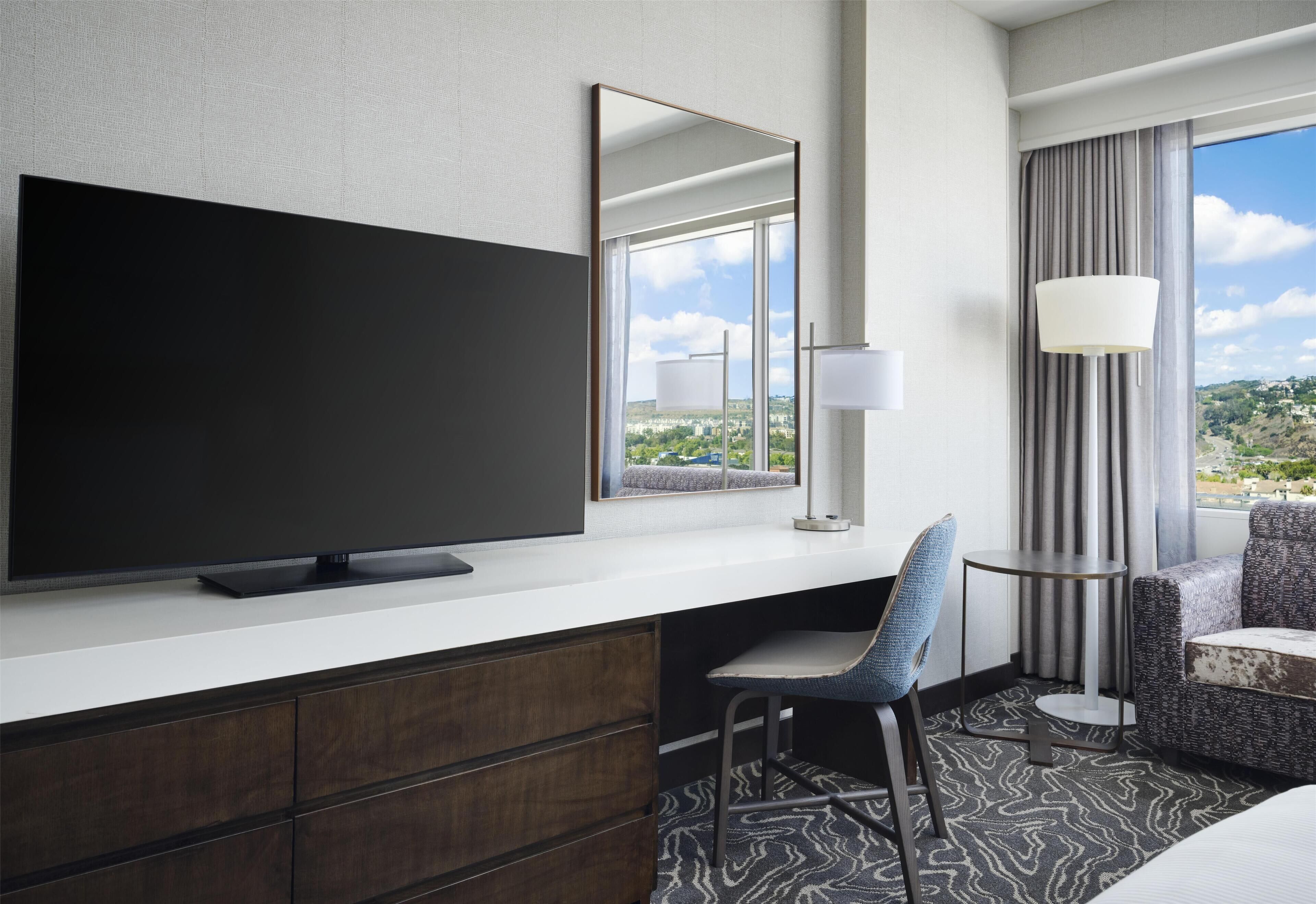 A hotel room with a large flat-screen TV on a white console, a chair, a small table, and a view of the city through the window.
