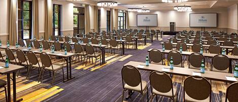 A large conference room with rows of tables and chairs, water bottles on each table, and a projector screen displaying the Hilton logo.