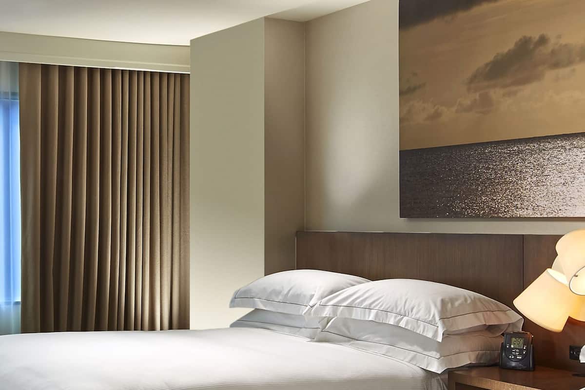 A hotel room with a bed, curtains, a painting, and a nightstand with a lamp and clock.