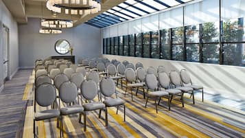 Un salón de conferencias con filas de sillas, un ventanal con vistas a árboles y una araña.