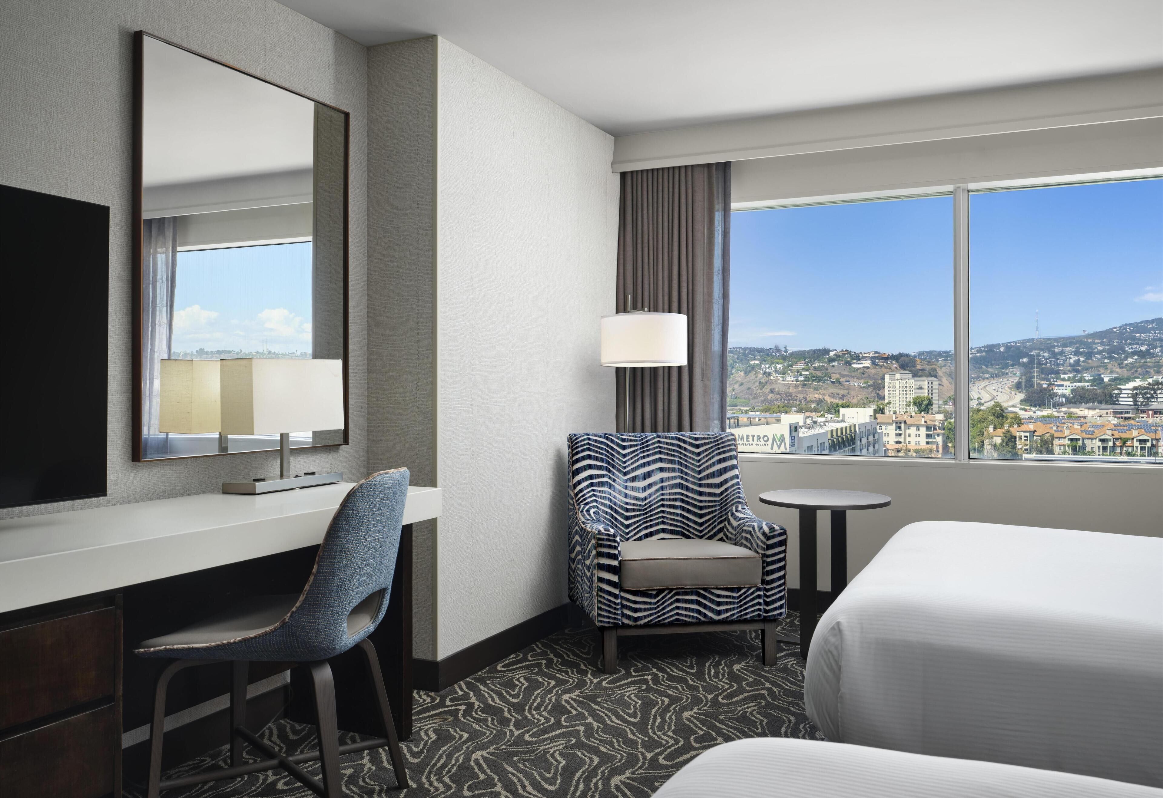 Ein Hotelzimmer mit einem Bett, einem Schreibtisch, einem Sessel, einem kleinen Tisch, einer Lampe und einem großen Fenster mit Blick auf die Stadt.