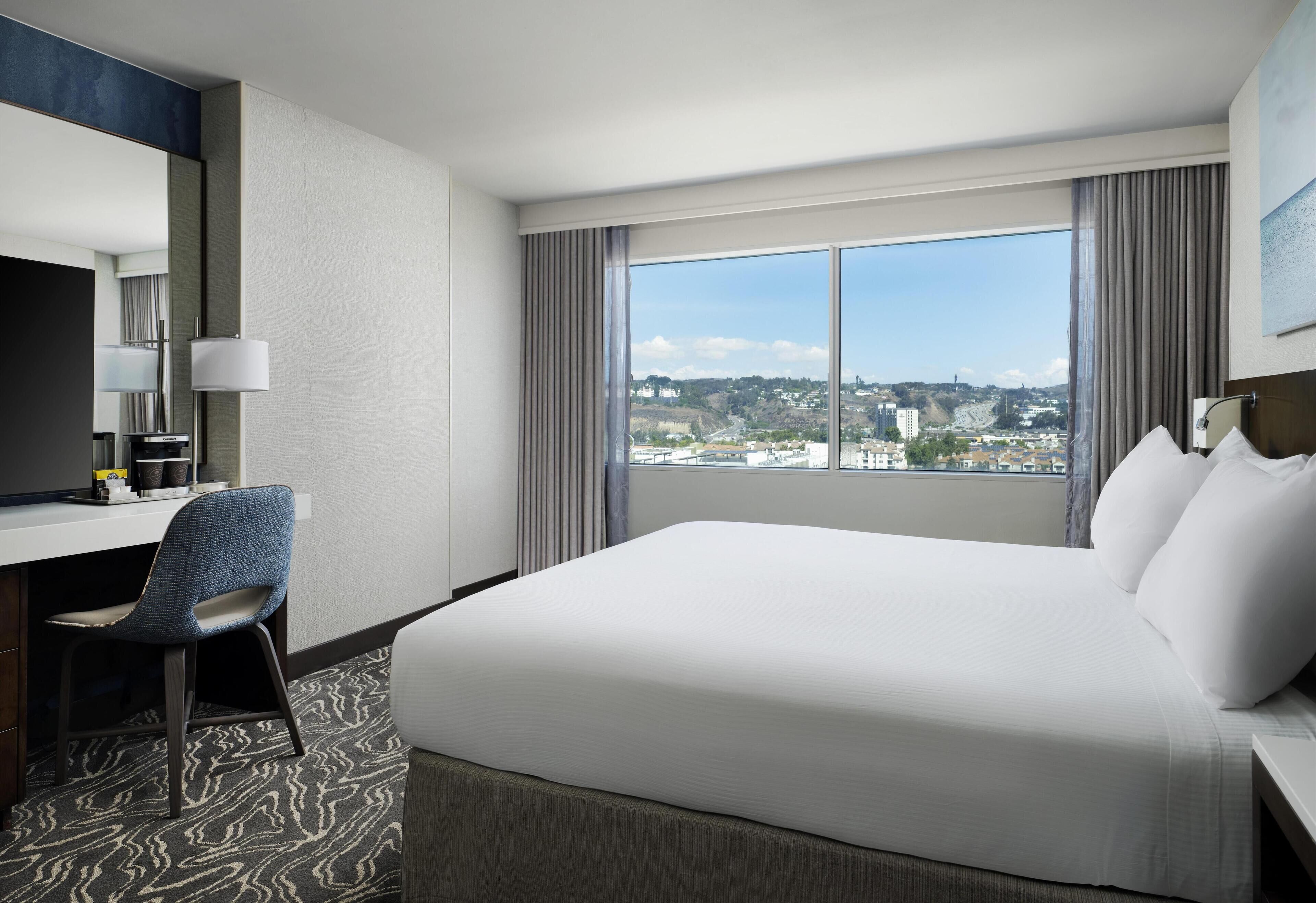 A hotel room with a large bed, a desk with a chair, a view of the city through large windows, and a modern design.