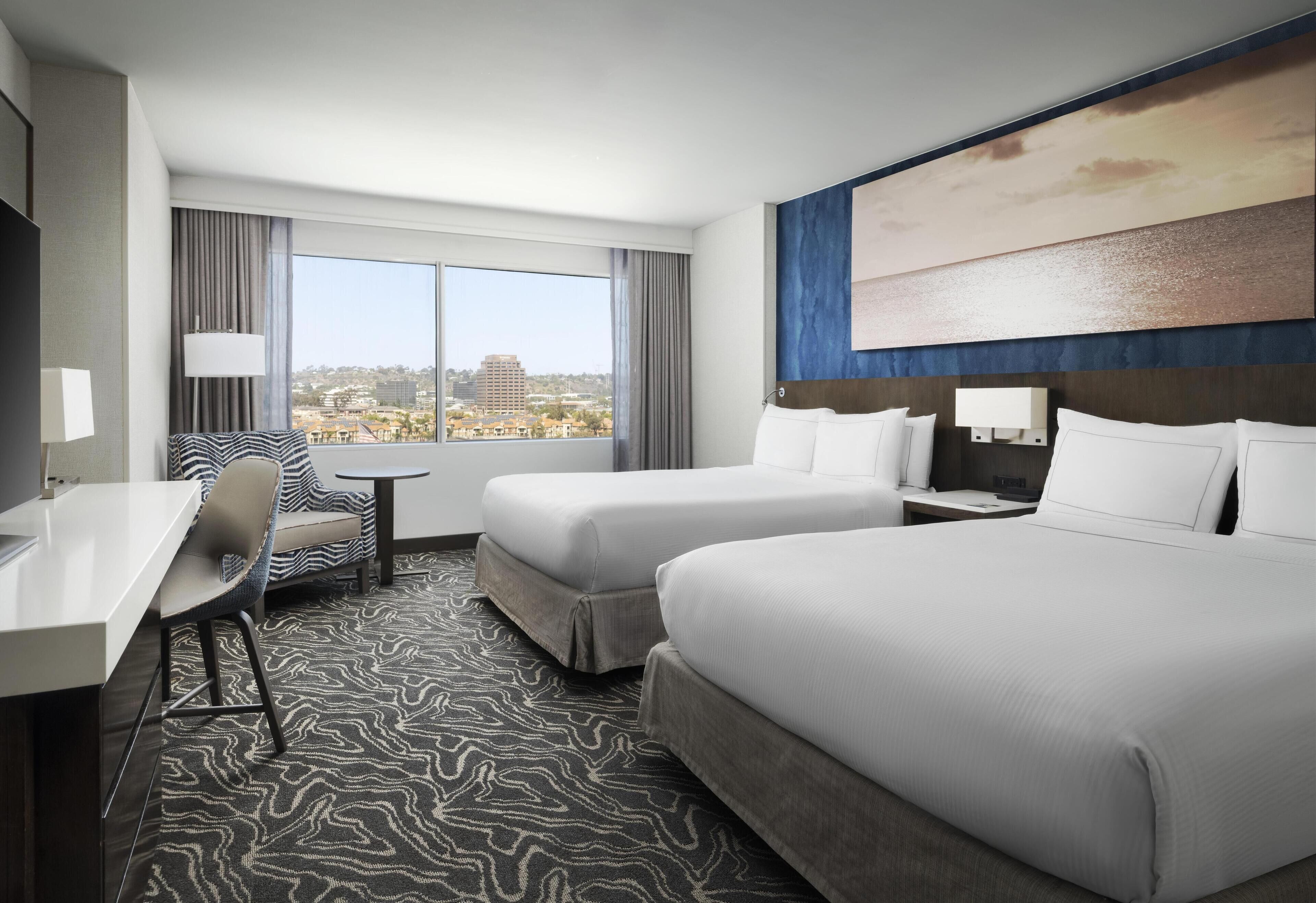 A hotel room with two beds, a desk, a chair, a small table, and a large window offering a city view.