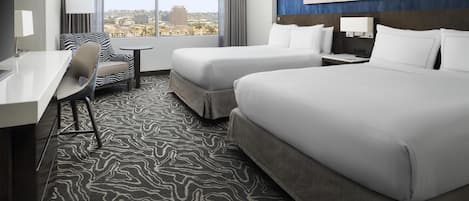 A hotel room with two beds, a desk, a chair, a small table, and a large window offering a city view.