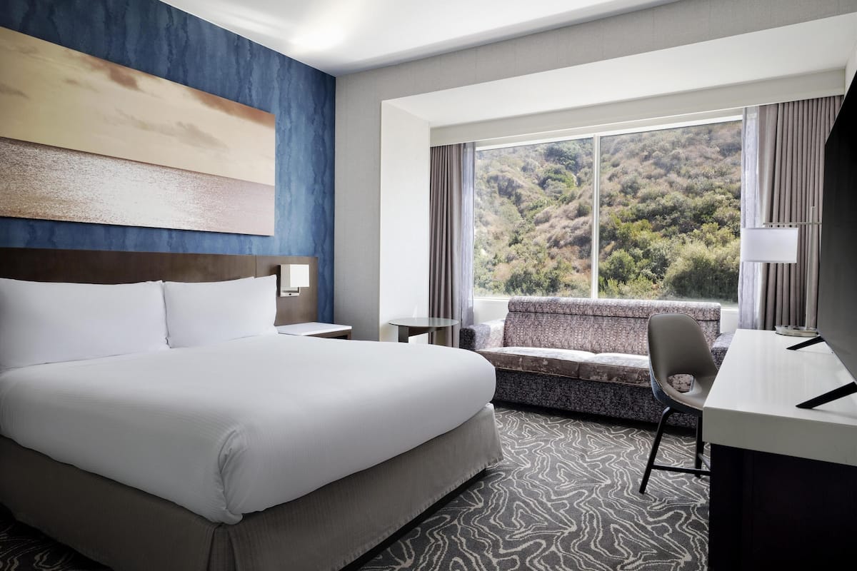A hotel room with a large bed, a desk, a chair, a window with a view of a forest, and a painting on the wall.