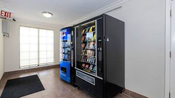 Quarto com uma máquina de vendas preta e uma máquina de Pepsi azul, janela com persianas e uma porta branca.