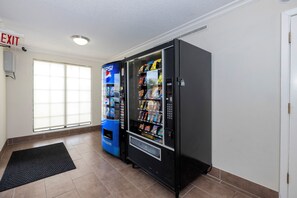 Una habitación con una máquina expendedora negra y una máquina Pepsi azul, una ventana con persianas y una puerta blanca.