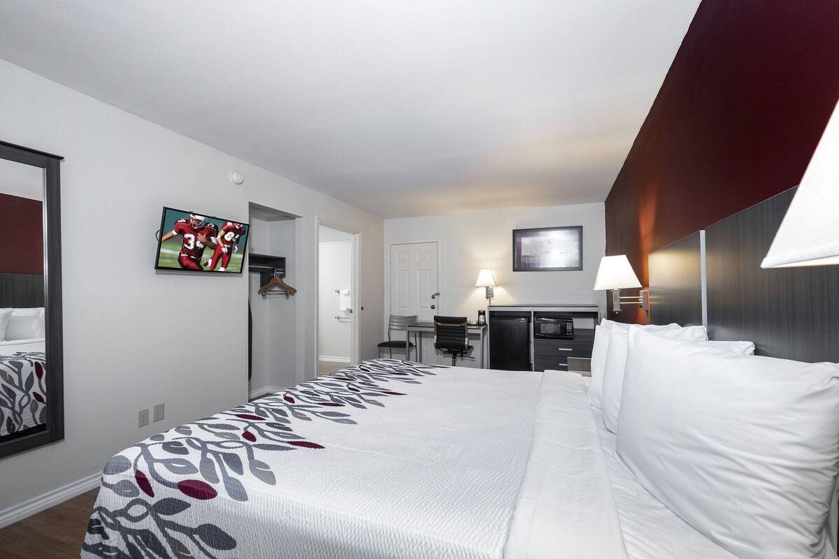 A hotel room with a large bed, a desk, a chair, a TV, and a kitchenette.