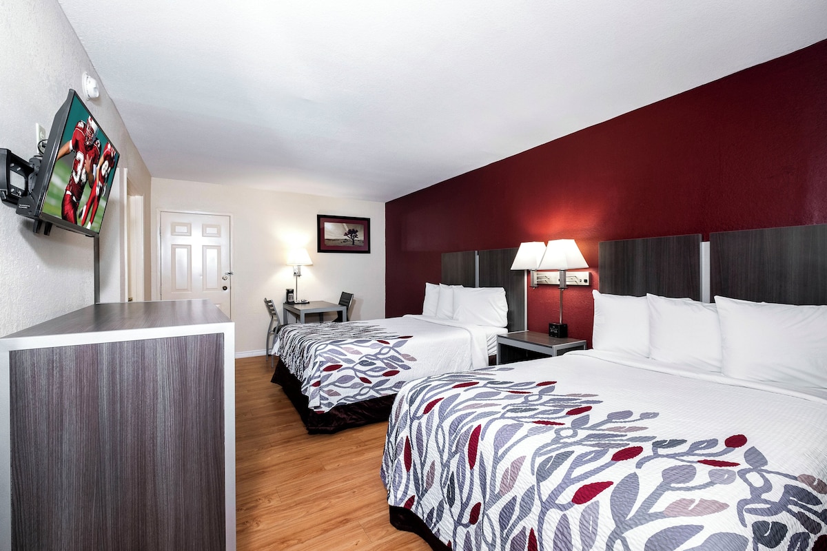 Quarto de hotel com duas camas, uma escrivaninha, uma cadeira, televisão na parede e abajur na mesa de cabeceira.