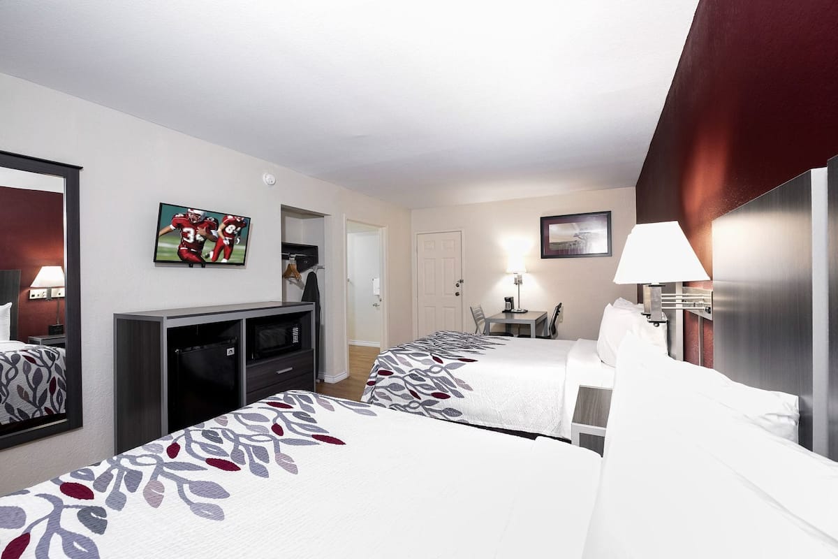 A hotel room with two beds, a TV, a desk with a chair, a lamp, and a door leading to another room.