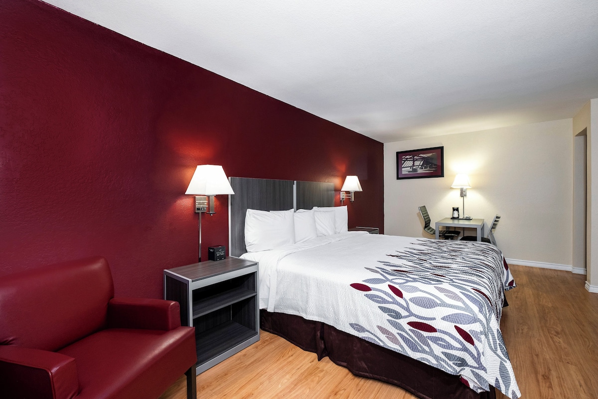 Quarto de hotel com cama, escrivaninha, cadeira e sofá vermelho.
