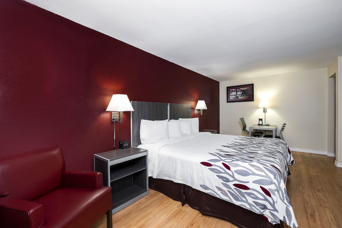 A hotel room with a red wall, a bed with a patterned bedspread, a red sofa, a small table with a lamp, and a chair.