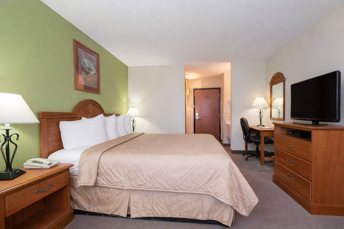 A hotel room with a bed, bedside tables, a desk with a chair, a television, and a door leading to the bathroom.