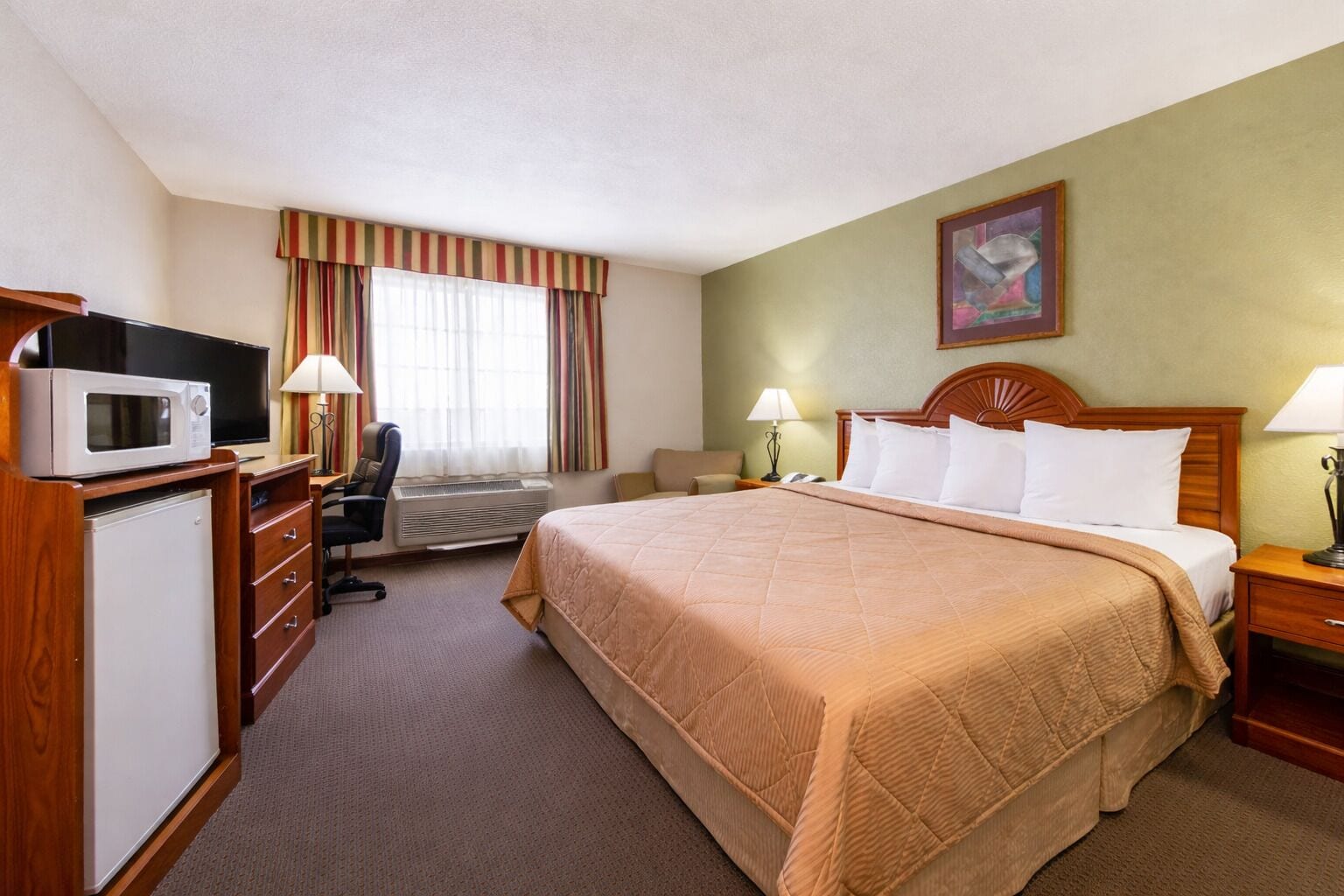 Une chambre d’hôtel avec un lit, un bureau avec un ordinateur, un four à micro-ondes, une chaise, une table de chevet et un tableau au mur.