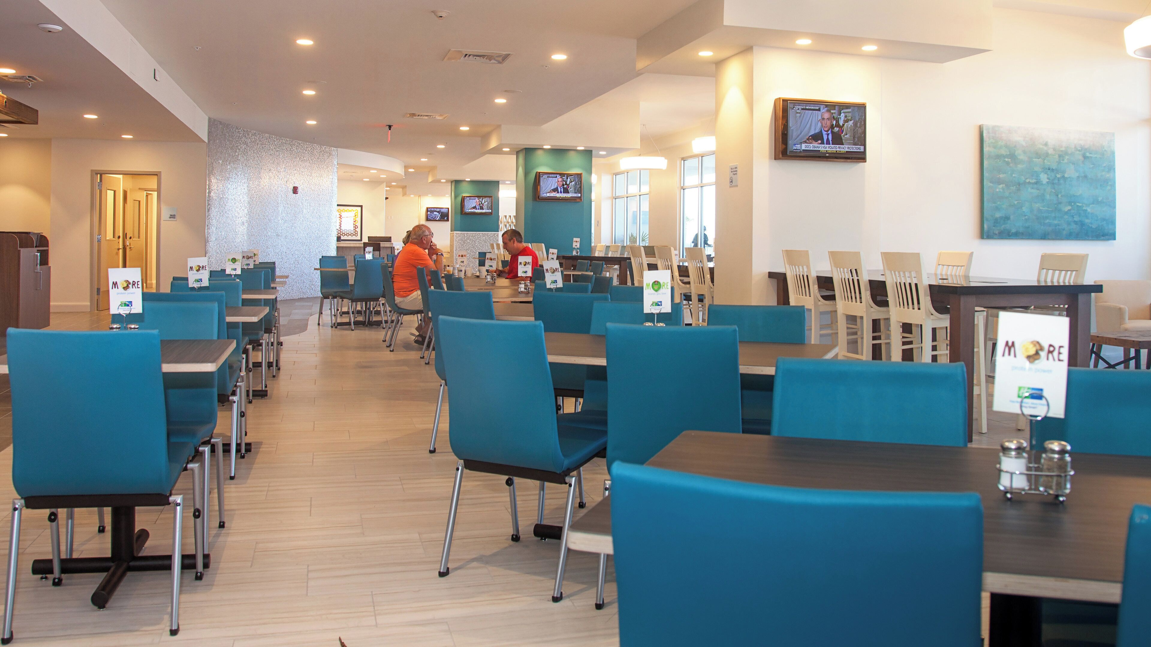 A hotel dining area with multiple tables and chairs, a television mounted on the wall, and a sign indicating the restaurant's name.