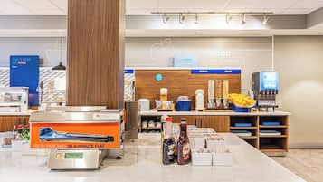 Un área de desayuno con una máquina para hacer panqueques, dispensadores de bebidas y una exhibición de frutas.