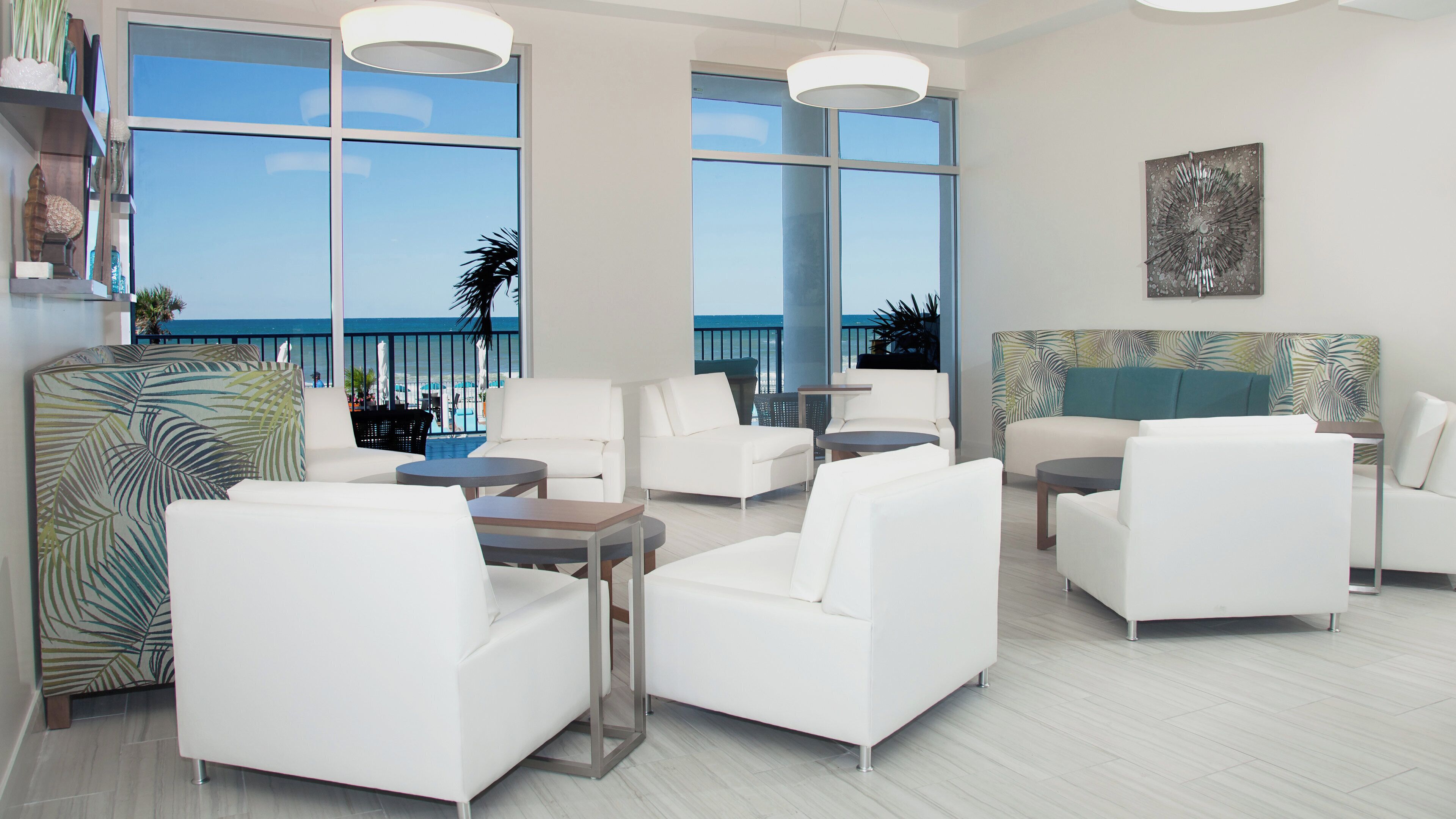 A modern lounge area with white furniture, large windows with a view of the beach, and a tropical-themed wall.