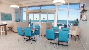 Un espace de restauration moderne avec des chaises et des tables de couleur turquoise, de grandes fenêtres donnant sur l’océan et un balcon.
