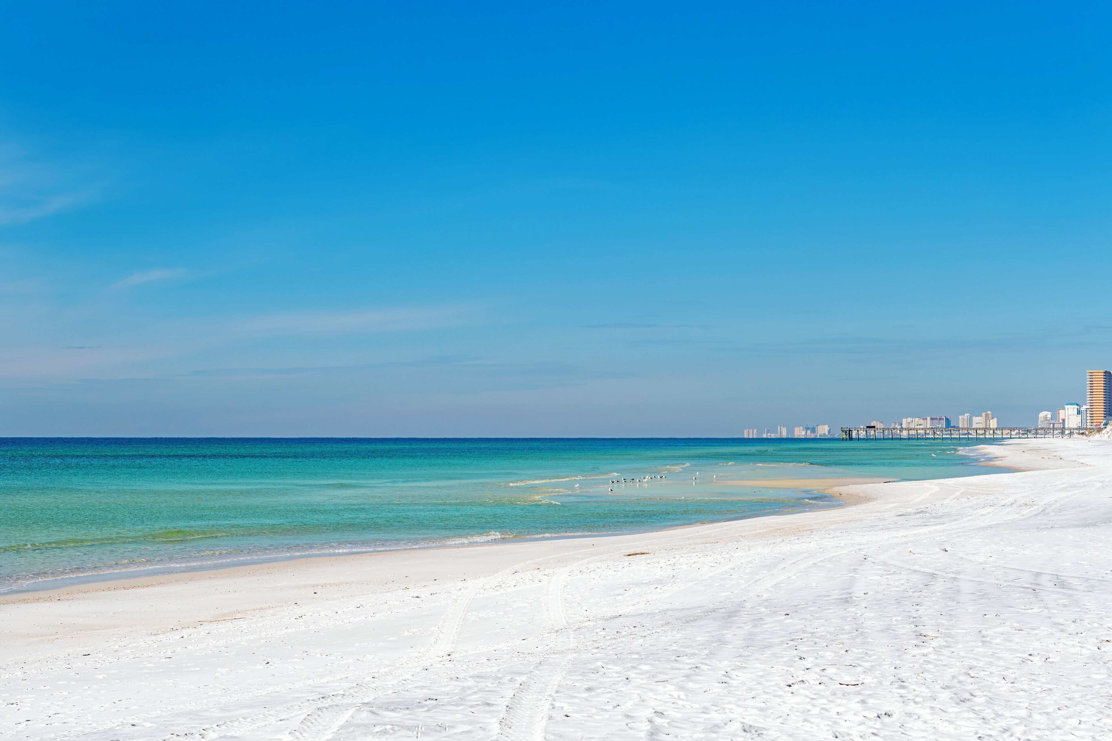 A wide sandy beach with clear turquoise waters and a distant city skyline.