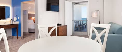 A modern hotel room with a dining area, a balcony with chairs, and a bed with a nightstand.