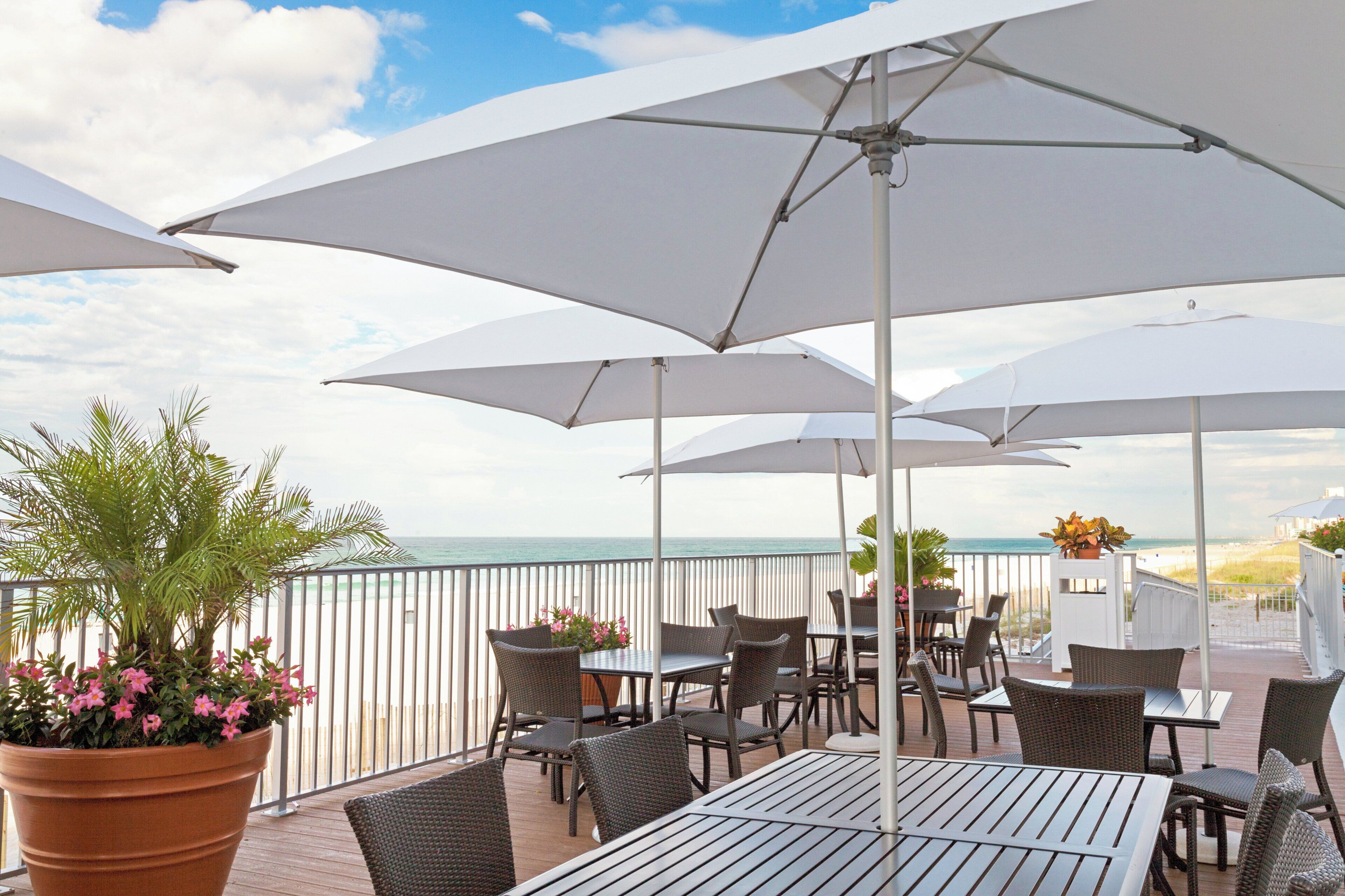Un patio exterior con mesas y sillas, sombrillas blancas y vista al océano.