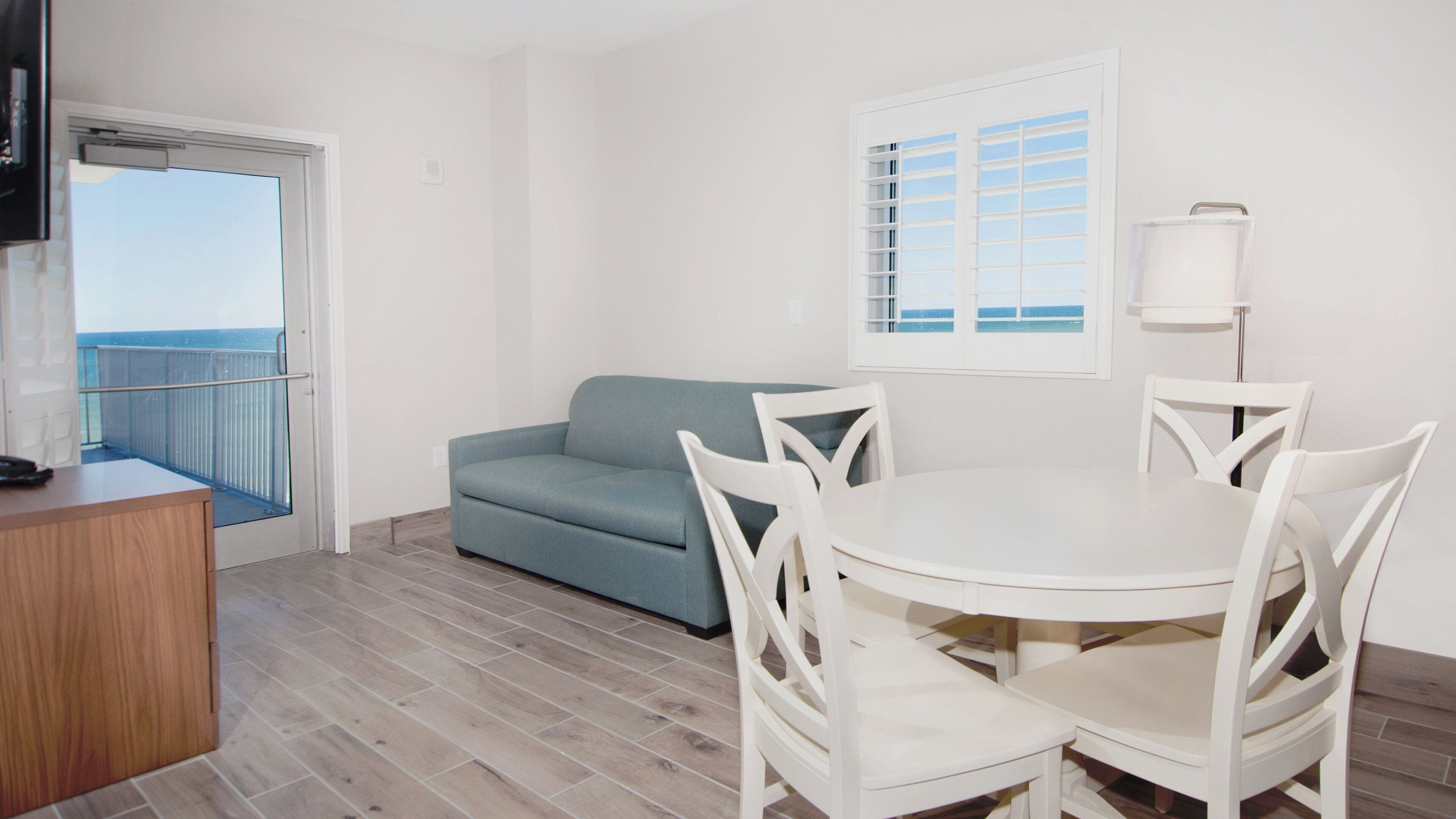 A room with a balcony, a sofa, a dining table with chairs, and a television.