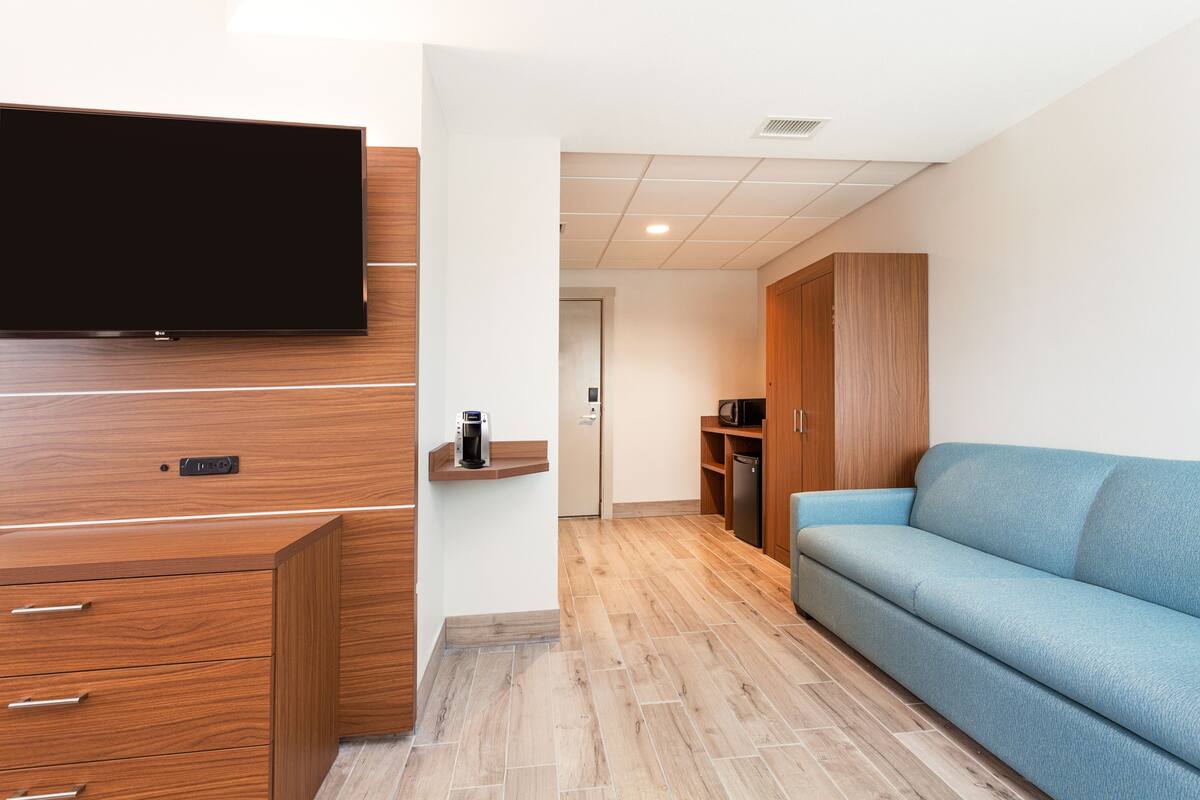 Un salon moderne avec un téléviseur à écran plat fixé à un meuble en bois, un canapé bleu et un coin cuisine doté de meubles en bois et d’un four à micro-ondes.