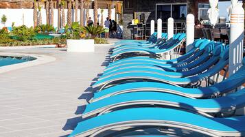 A row of sun loungers by a poolside, with a stone pathway and palm trees in the background.