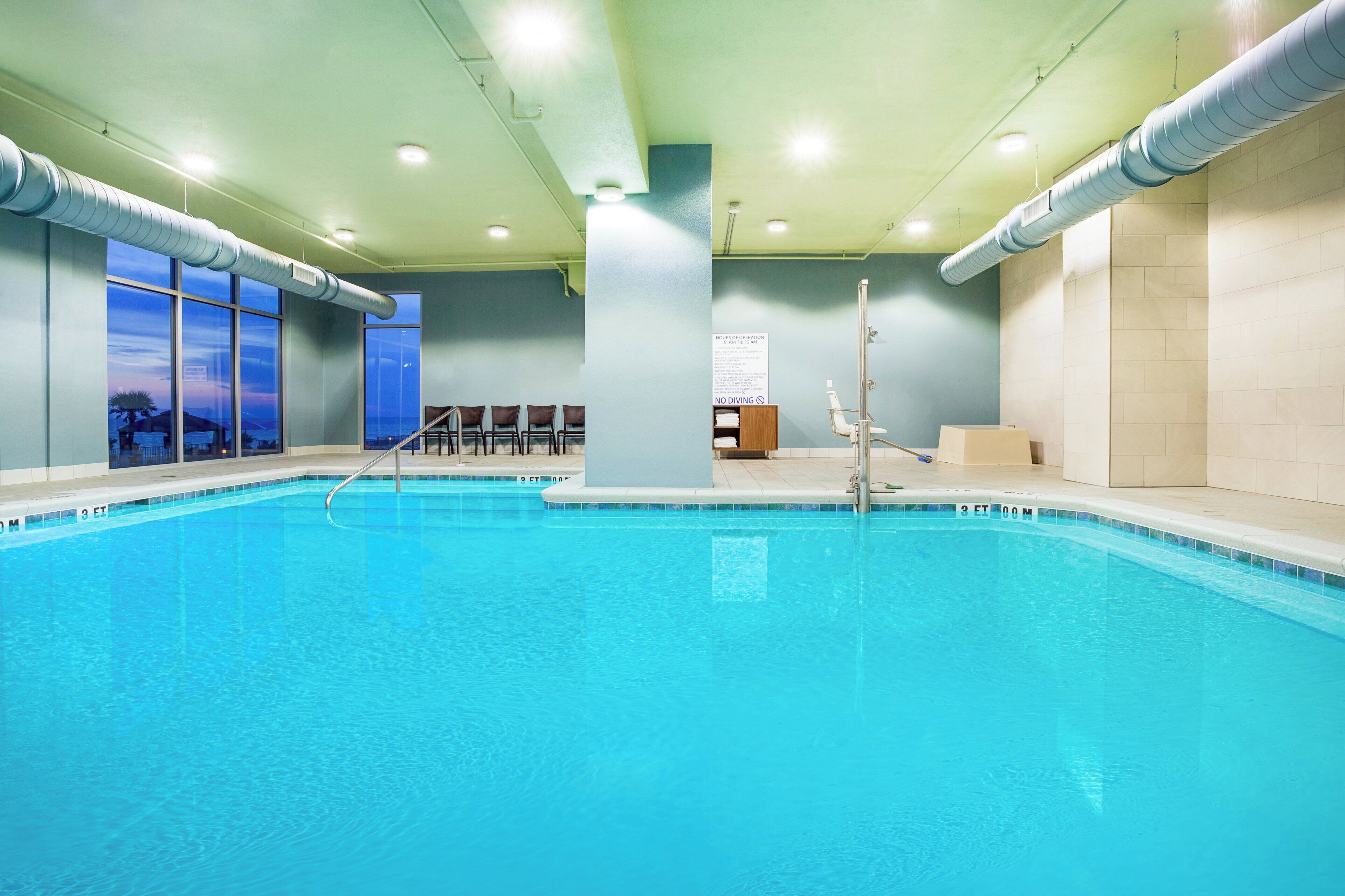 An indoor swimming pool with clear blue water, a changing room area, and a view of the outside through large windows.