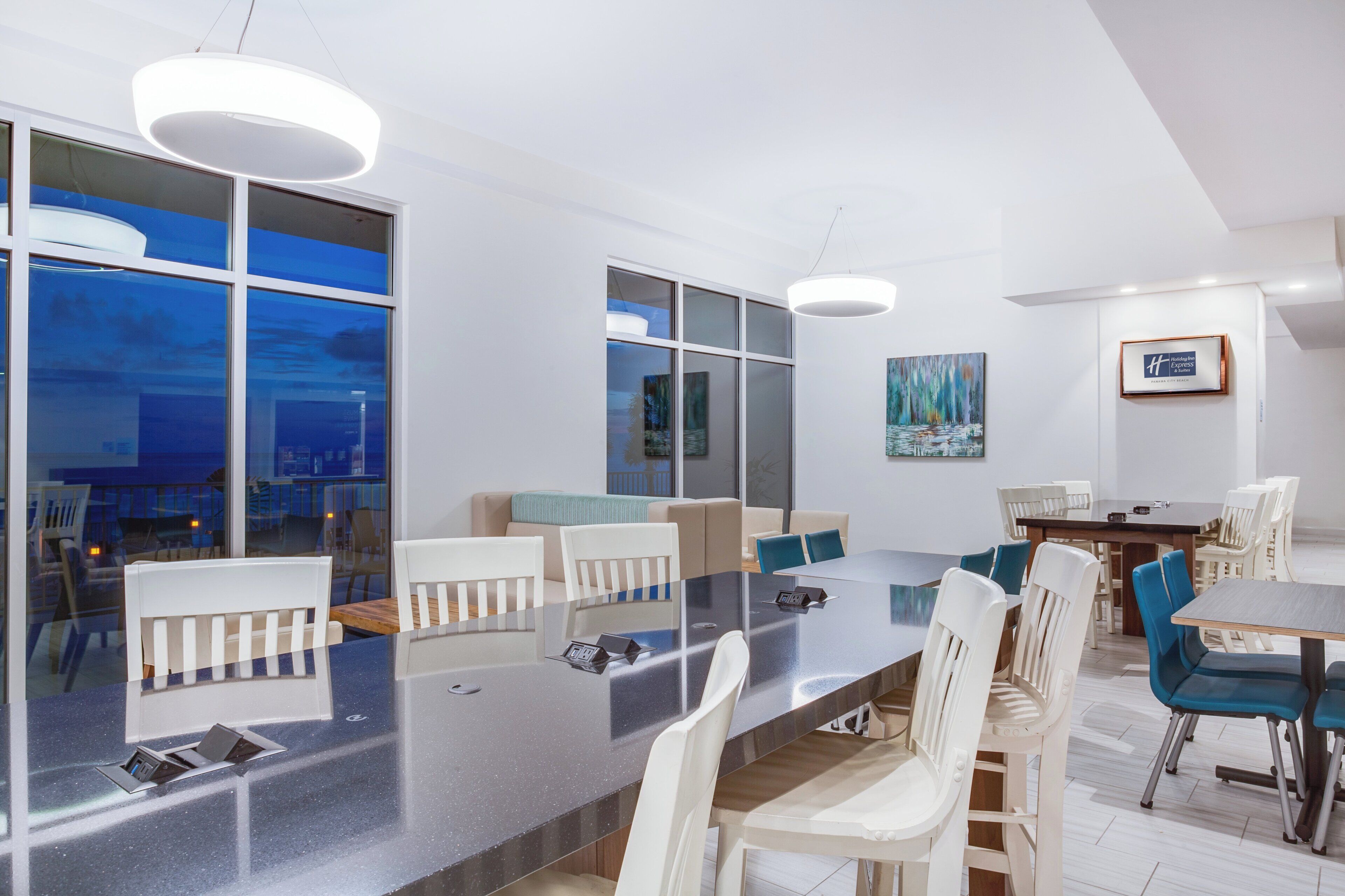 Una habitación de hotel moderna con un amplio comedor, varias sillas, un sofá y un ventanal con vistas a la ciudad.