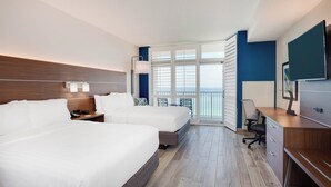 Ein Hotelzimmer mit zwei Betten, einem Schreibtisch, einem Stuhl und einem großen Fenster mit Blick auf den Ozean.