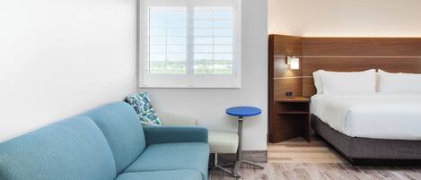Quarto de hotel com sofá azul, uma mesa redonda pequena, cama com roupa de cama branca e janela com persianas.
